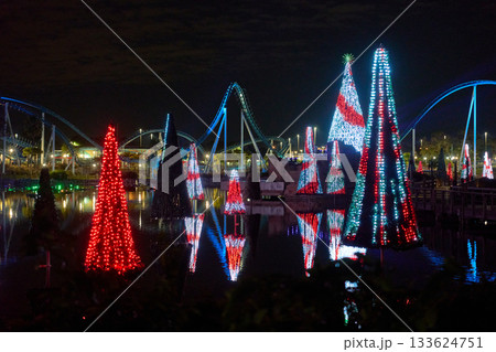 Striped red and white Christmas light trees float on a lagoon with vivid reflections. Curved coaster tracks and park lights create a bright winter night panorama. Striped red and white Christmas light trees float on a lagoon with vivid reflections. Curved coaster tracks and park lights create a bright winter night panorama. 133624751