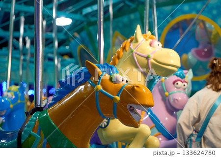 Close view of a bright cartoon carousel horse with a blue mane and shiny bridle. Vibrant lights and other ride figures glow in the background. 133624780