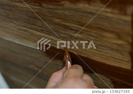 A close-up view of a person's hand using a putty knife to apply brown grout between the joints of wood-look ceramic floor tiles. The man is doing DIY home improvement and renovation work. 133627193