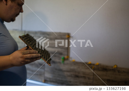 Laying tiles on a wall in a house A man is renovating a house. He holds ceramic tiles with adhesive applied, preparing to install them on the wall during the renovation, focusing on the precise work. 133627198