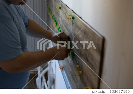 Man using tile leveling tools to install wood-look ceramic wall tiles in a bathroom renovation, close-up of skilled hands ensuring precise, even tile placement and finish. 133627199