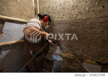 A worker in protective clothing operates a hammer drill to remove old plaster and wall tiles. A man uses a hammer drill for demolition work during a home or bathroom renovation project. A worker in protective clothing operates a hammer drill to remove old plaster and wall tiles. A man uses a hammer drill for demolition work during a home or bathroom renovation project. 133627200