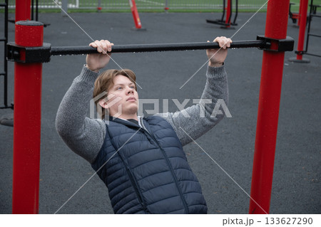 Young man performing pull-up exercise at an outdoor gym, holding onto a bar. Strength training in public fitness area. Outdoors 133627290