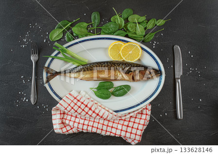 Whole smoked mackerel fish on white oval plate, gray black stone like table under, rock salt, lemon slices and green leaves salad near. View from above 133628146
