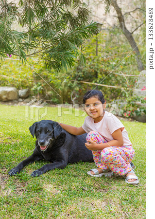 A young girl is sitting on the grass next to a black dog A young girl is sitting on the grass next to a black dog 133628489