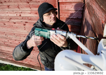 Drilling passage for surveillance camera cable, installer guides power drill into wooden wall of rural house, technician bores opening through facade. Drilling passage for surveillance camera cable, installer guides power drill into wooden wall of rural house, technician bores opening through facade. 133629692