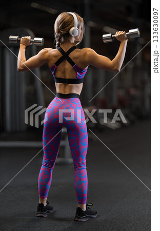 In a gym setting a girl focuses on her form as she lifts dumbbells for shoulder exercises. She wears a colorful workout outfit and headphones showing dedication. In a gym setting a girl focuses on her form as she lifts dumbbells for shoulder exercises. She wears a colorful workout outfit and headphones showing dedication. 133630097