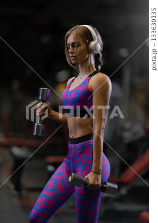girl focuses on her form while doing bicep curls with shiny dumbbells. She wears workout clothes and headphones in a well lit gym showing determination and strength. 133630135