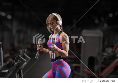 girl in activewear lifts shiny dumbbells for bicep curls in a gym. She wears headphones and concentrates on her exercise showcasing strength and determination during her workout. girl in activewear lifts shiny dumbbells for bicep curls in a gym. She wears headphones and concentrates on her exercise showcasing strength and determination during her workout. 133630148