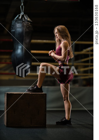 girl stands on a cube in a boxing gym focusing on her balance and strength. The surrounding area features a boxing bag and training equipment. 133630168