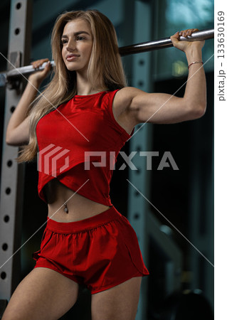 A girl performs a squat with a barbell in a gym showcasing her strength and determination. She wears a bright red outfit and appears focused on her workout routine. A girl performs a squat with a barbell in a gym showcasing her strength and determination. She wears a bright red outfit and appears focused on her workout routine. 133630169