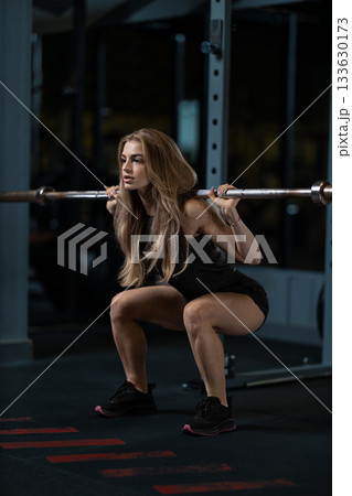 Girl is performing a barbell squat in a gym setting. She focuses on her form while lifting weights in an effort to enhance her strength and fitness level. 133630173