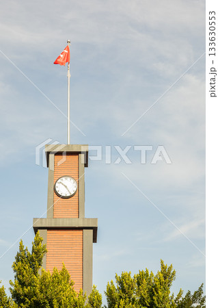 Brick clock tower with flag against blue sky surrounded by trees Brick clock tower with flag against blue sky surrounded by trees 133630553