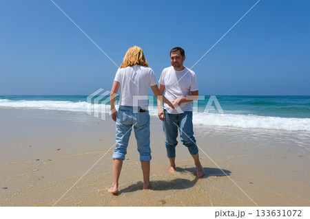Joyful couple playing at the water's edge on a sunny beach, dressed in casual jeans and white tshirts 133631072