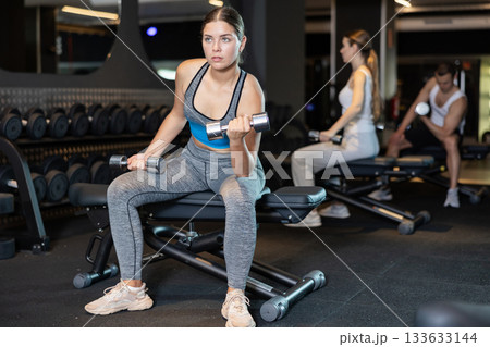 Determined young woman working out with dumbbell in sports hall Determined young woman working out with dumbbell in sports hall 133633144
