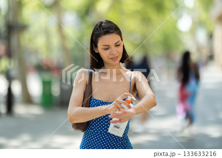 Young brunette girl applying sunscreen to her skin while walking in city Young brunette girl applying sunscreen to her skin while walking in city 133633216