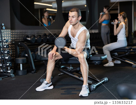 Determined young man working out with dumbbell in sports hall Determined young man working out with dumbbell in sports hall 133633217