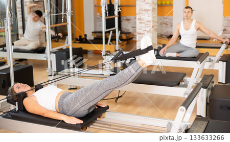 Girl works out in gym and performs set of exercises to stretch leg muscles with help of reformer Girl works out in gym and performs set of exercises to stretch leg muscles with help of reformer 133633266