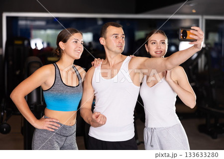 Female gym visitors with man coach takes pictures of herself on phone, takes selfie in gym Female gym visitors with man coach takes pictures of herself on phone, takes selfie in gym 133633280