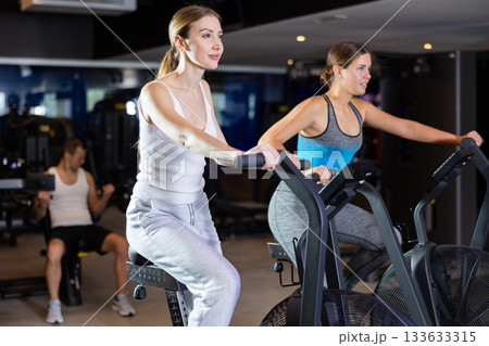 Girl performs exercises with stationary air bicycle in gym Girl performs exercises with stationary air bicycle in gym 133633315