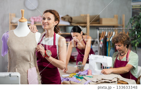 Female tailor sews new dress on mannequin in sewing studio. Man works on sewing machine. Another tailor draws sketch of dress Female tailor sews new dress on mannequin in sewing studio. Man works on sewing machine. Another tailor draws sketch of dress 133633348
