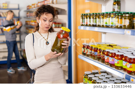 In Russian goods store, woman choose jar of pickled tomatoes and cucumbers 133633446
