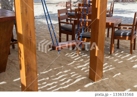 Tranquil beach scene with wooden swing and seaside dining area in soft sand 133633568