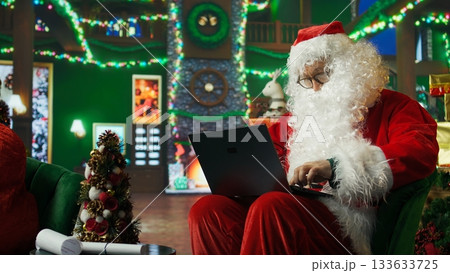 Santa Claus performer typing on laptop, preparing shipping parcel with gift to be sent to kids. Man dressed as Father Christmas scheduling goods delivery, preparing to send presents to children 133633725