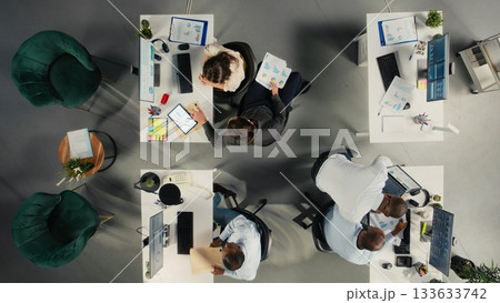 Top down view of corporate employees study business reports and analytics. Colleagues share insights on forecasting, documentation, and targets to improve growth strategy. 133633742