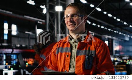Engineer oversees assembly tools on tablet in industry plant, surrounded by metal and steel machinery within the factory. Strong workforce involvement and heavy manufacturing tasks. Engineer oversees assembly tools on tablet in industry plant, surrounded by metal and steel machinery within the factory. Strong workforce involvement and heavy manufacturing tasks. 133633789