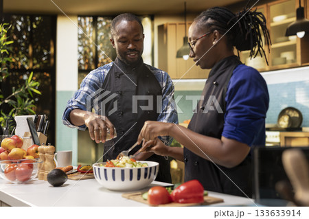African american man seasoning the salad with mix of spices from a jar, pouring condiments for a tasty vegetarian meal. Smiling woman mixing fresh vegetables together, healthy lifestyle. African american man seasoning the salad with mix of spices from a jar, pouring condiments for a tasty vegetarian meal. Smiling woman mixing fresh vegetables together, healthy lifestyle. 133633914