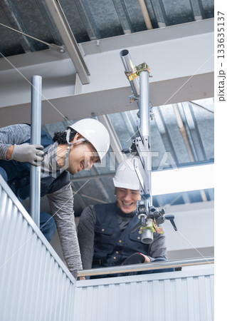 天井が近い高所の足場で単管パイプを固定するクランプなどの施工作業 鳶職や作業員のイメージ 笑顔で話す 天井が近い高所の足場で単管パイプを固定するクランプなどの施工作業 鳶職や作業員のイメージ 笑顔で話す 133635137