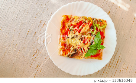 Slice of pizza with cheese, vegetables and basil on a white plate standing on a wooden table. Slice of pizza with cheese, vegetables and basil on a white plate standing on a wooden table. 133636349