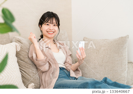 リビングのソファーに座りながらスマホのアプリを使うアジア人女性（ガッツポーズ） 133640667