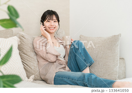 ホットドリンクを飲みながらスマホで電話する日本人女性(家,リビング,笑顔) ホットドリンクを飲みながらスマホで電話する日本人女性(家,リビング,笑顔) 133641050