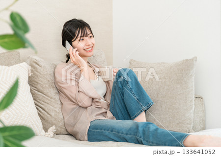 ホットドリンクを飲みながらスマホで電話する日本人女性(家,リビング,笑顔) ホットドリンクを飲みながらスマホで電話する日本人女性(家,リビング,笑顔) 133641052