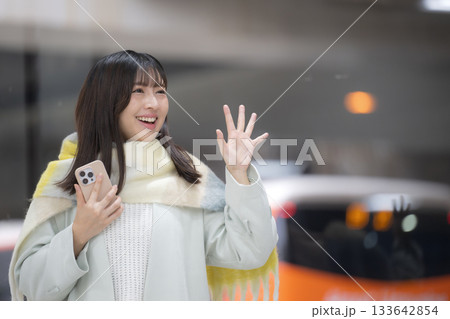 冬の駅のホームやバスターミナルなどでスマホを持つかわいいストールを巻いた女性、手を振るバイバイのポー 133642854