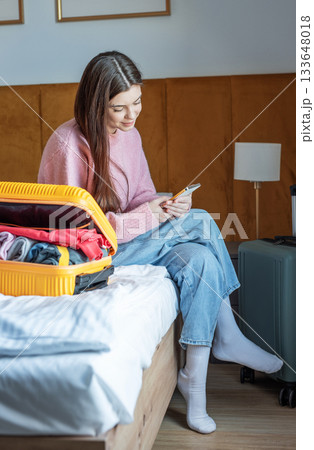 Woman using phone near luggage in hotel room 133648018