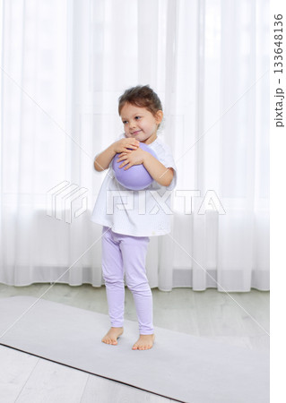 Toddler girl embracing purple pilates ball, standing on a yoga mat in a bright room 133648136