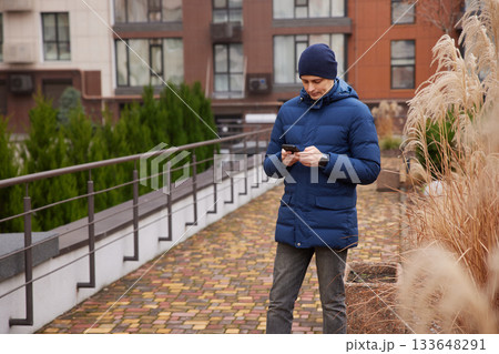 Man in winter coat and beanie texting on phone outdoors near ornamental grass Man in winter coat and beanie texting on phone outdoors near ornamental grass 133648291