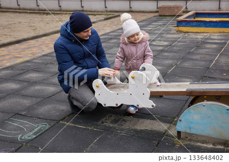 Father playing with toddler daughter on a seesaw at an outdoor playground in winter Father playing with toddler daughter on a seesaw at an outdoor playground in winter 133648402