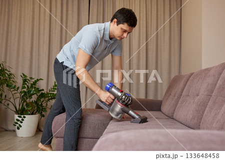 Man vacuuming sofa in living room, performing housework for maintaining home cleanliness Man vacuuming sofa in living room, performing housework for maintaining home cleanliness 133648458