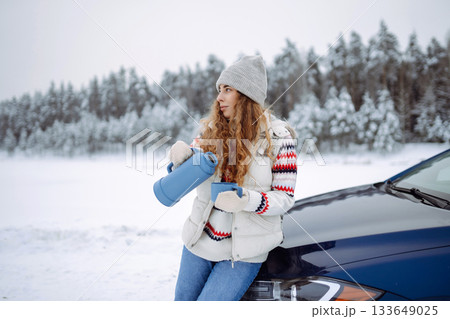 Attractive woman in a hat and gloves drinks a drink from a thermos near a car on a snowy road. Attractive woman in a hat and gloves drinks a drink from a thermos near a car on a snowy road. 133649025