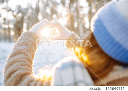 Woman in hat and scarf against backdrop of snowy winter forest holds hands in the shape of a heart. 133649315