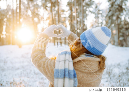 Woman in hat and scarf against backdrop of snowy winter forest holds hands in the shape of a heart. Woman in hat and scarf against backdrop of snowy winter forest holds hands in the shape of a heart. 133649316