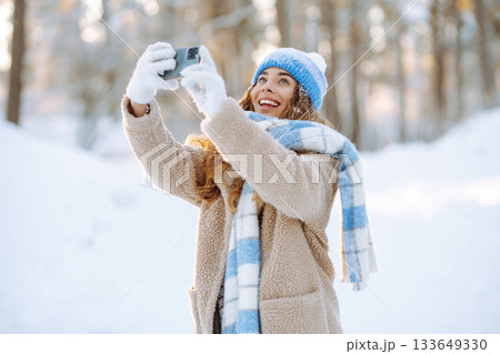 A young woman in a snowy park with a phone in her hands communicates via video and takes a selfie. 133649330