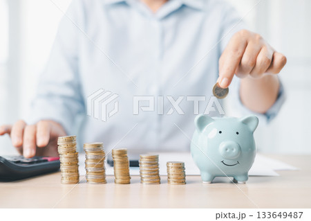 Concept of saving money and financial planning. A businesswoman puts coin into blue piggy bank on office desk with documents and laptop, symbolizing investment, budget management and future growth. 133649487