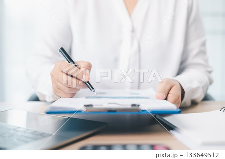 A businesswoman reviews financial report while holding documents and pointing with pen at office desk. Concept of accounting, budget management, financial analysis and corporate business planning. 133649512