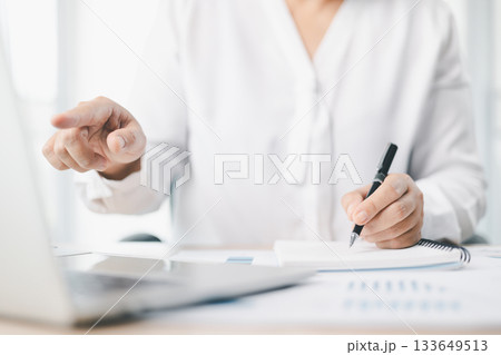 Concept of business planning and financial analysis. A businesswoman writes notes on documents with charts and reports, using laptop and calculator for budget management at modern office desk. 133649513