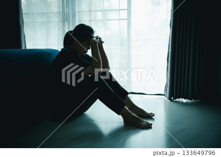 A lonely woman sits on the floor by window with hand on head in dark room. Concept of depression, sadness, anxiety, stress, mental health problem, loneliness and emotional struggle in life. 133649796